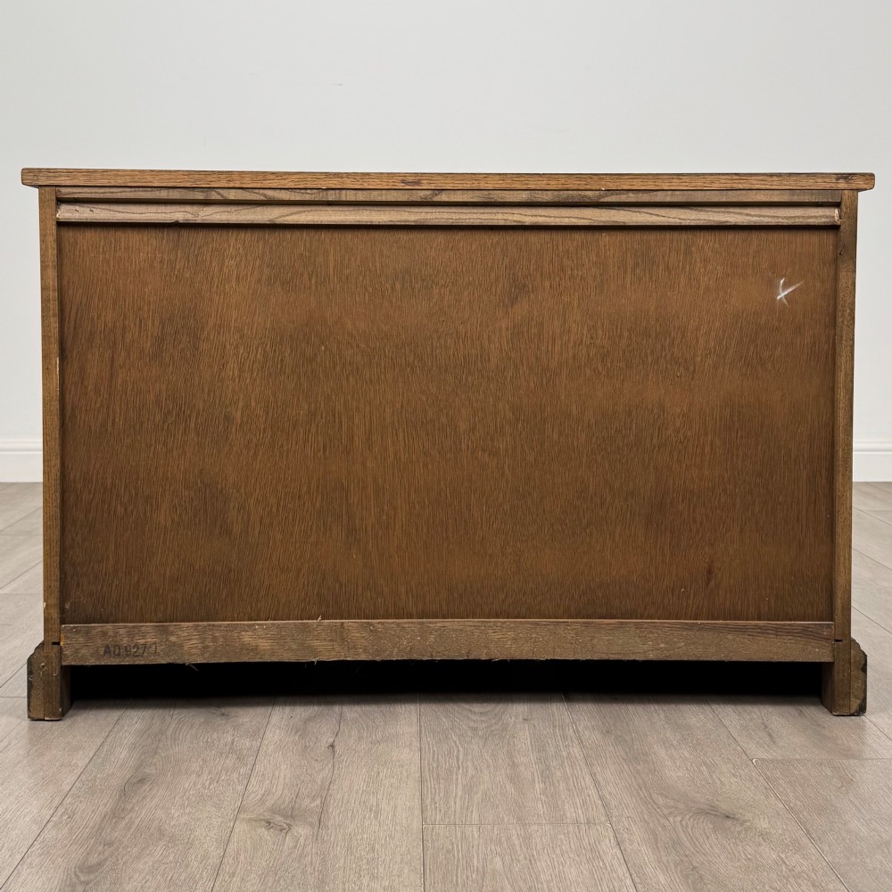 A dark oak low cupboard featuring canted corners, carved linenfold door panels, an upper drawer with carved foliage, and brass hinges. View VI