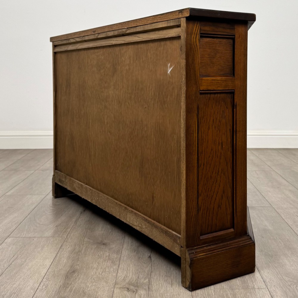 A dark oak low cupboard featuring canted corners, carved linenfold door panels, an upper drawer with carved foliage, and brass hinges. View VII