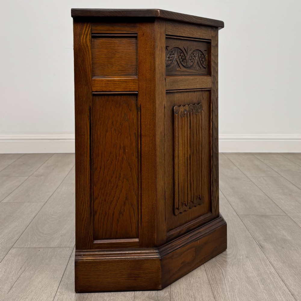 A dark oak low cupboard featuring canted corners, carved linenfold door panels, an upper drawer with carved foliage, and brass hinges. View VIII