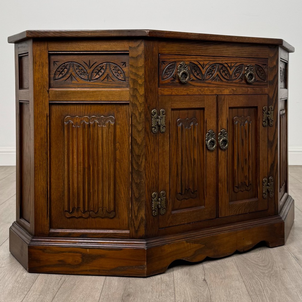 A dark oak low cupboard featuring canted corners, carved linenfold door panels, an upper drawer with carved foliage, and brass hinges. View IX