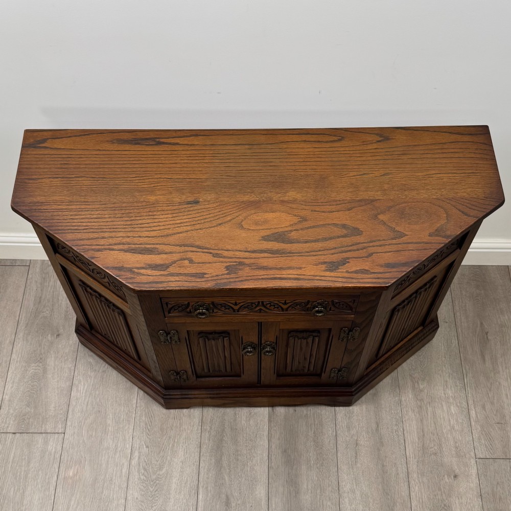 A dark oak low cupboard featuring canted corners, carved linenfold door panels, an upper drawer with carved foliage, and brass hinges. View X