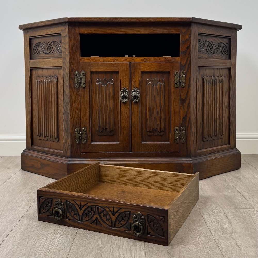 A dark oak low cupboard featuring canted corners, carved linenfold door panels, an upper drawer with carved foliage, and brass hinges. View XI