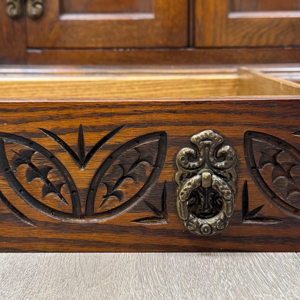 A dark oak low cupboard featuring canted corners, carved linenfold door panels, an upper drawer with carved foliage, and brass hinges. View XII