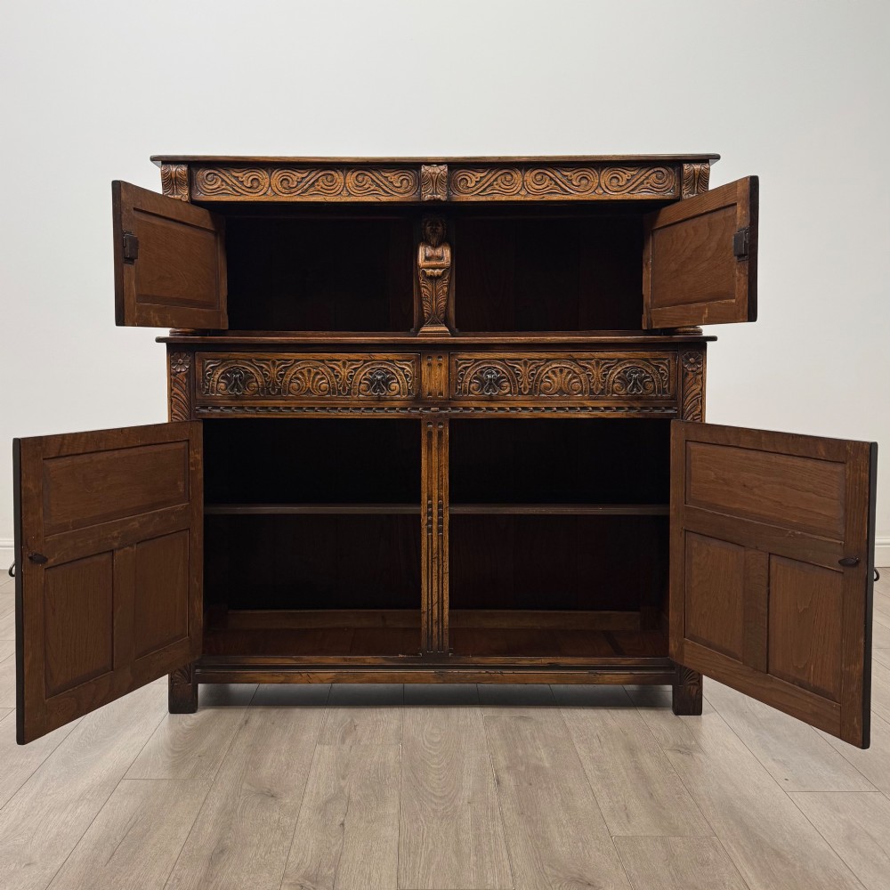 A dark carved oak court cupboard featuring two top doors with relief carving, bulbous turned supports, two drawers, and two panelled cupboard doors below. View II