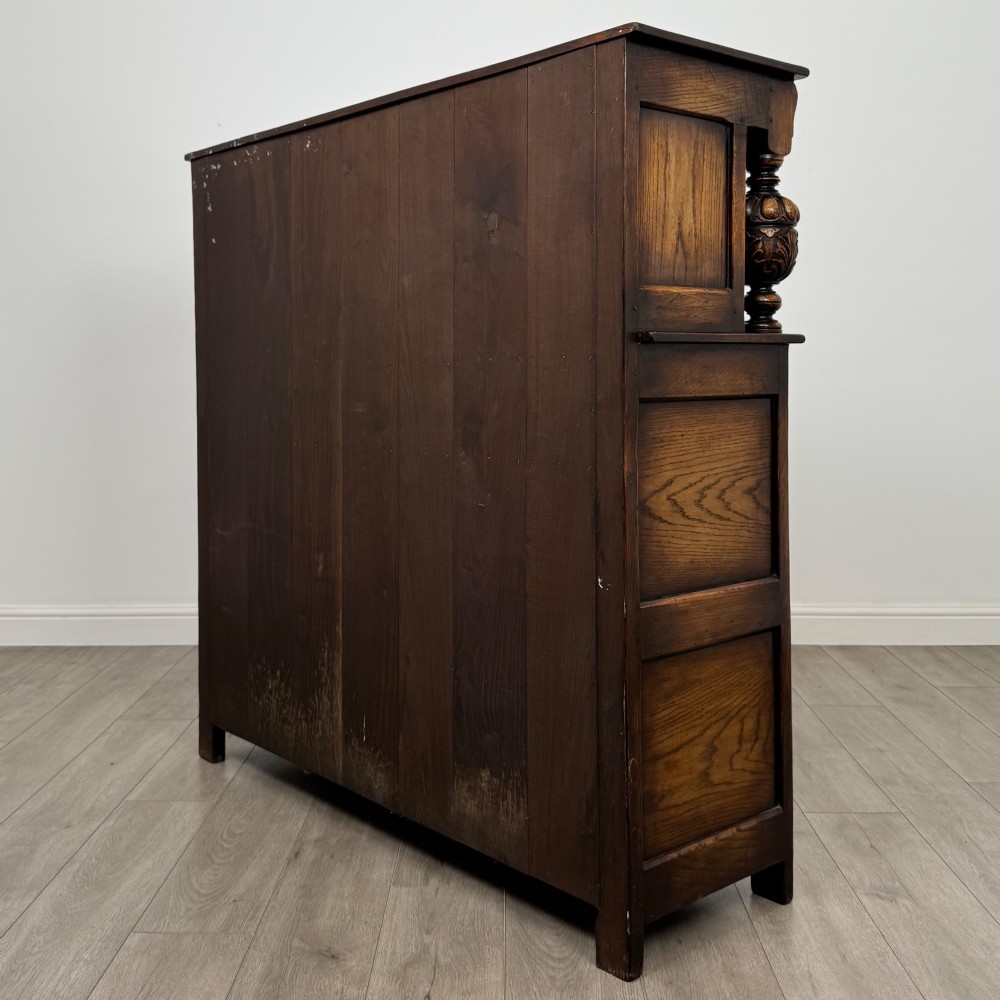 A dark carved oak court cupboard featuring two top doors with relief carving, bulbous turned supports, two drawers, and two panelled cupboard doors below. View VII