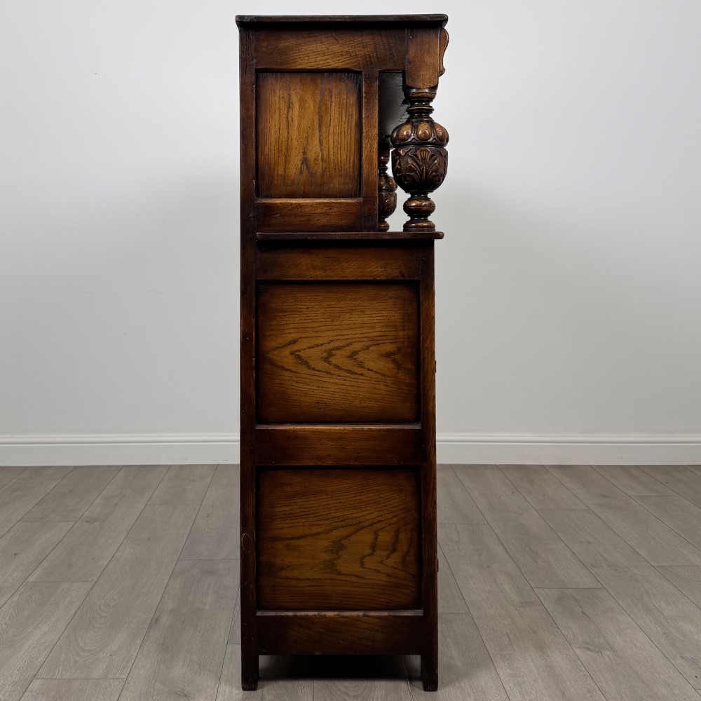 A dark carved oak court cupboard featuring two top doors with relief carving, bulbous turned supports, two drawers, and two panelled cupboard doors below. View VIII