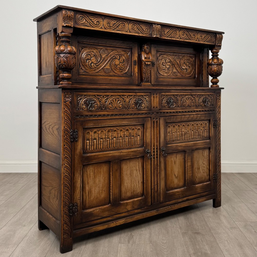 A dark carved oak court cupboard featuring two top doors with relief carving, bulbous turned supports, two drawers, and two panelled cupboard doors below. View IX