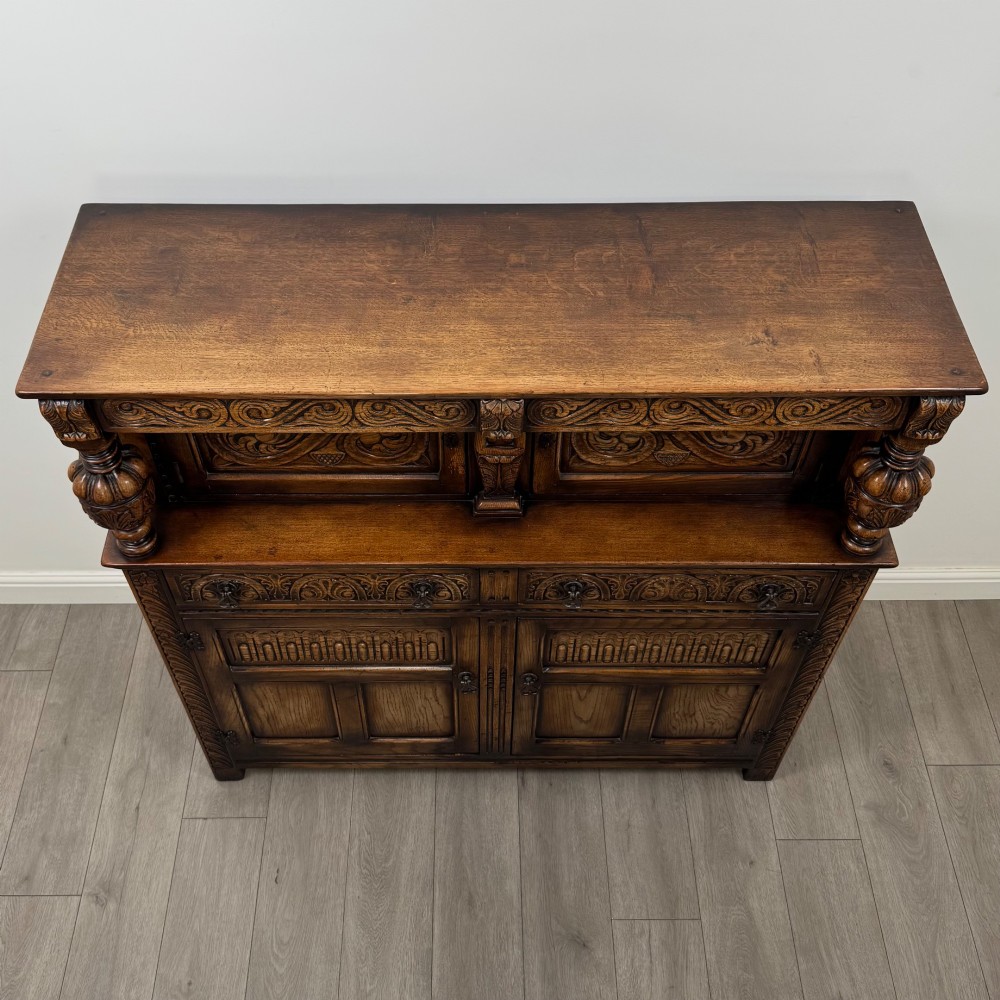 A dark carved oak court cupboard featuring two top doors with relief carving, bulbous turned supports, two drawers, and two panelled cupboard doors below. View X
