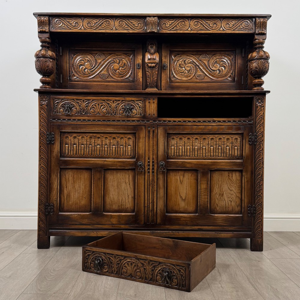 A dark carved oak court cupboard featuring two top doors with relief carving, bulbous turned supports, two drawers, and two panelled cupboard doors below. View XI