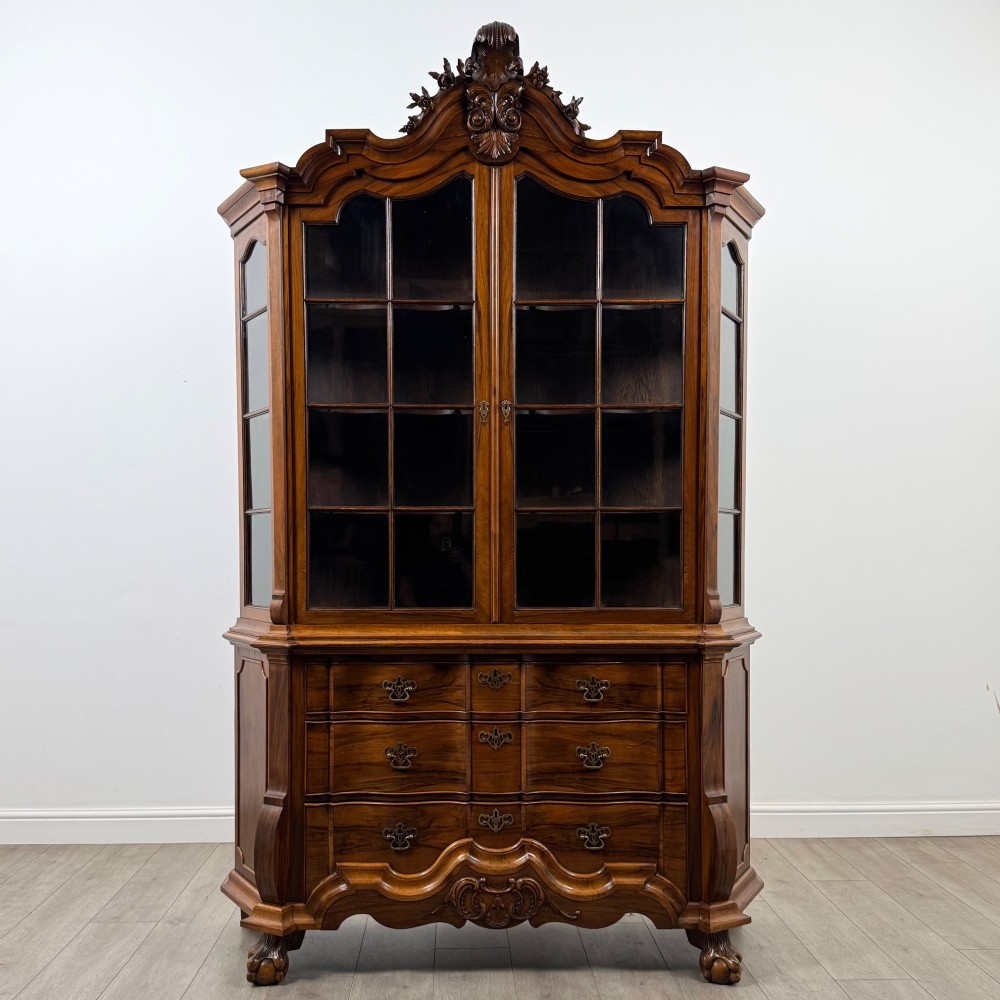 A Dutch walnut display cabinet featuring a carved bonnet-top pediment, multi-pane glazed doors, and a lower chest of drawers on carved claw-and-ball feet.
