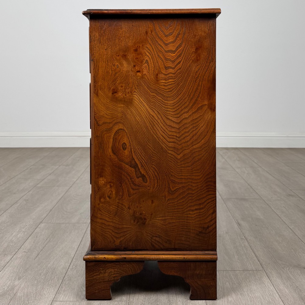 A small 20th-century serpentine chest of drawers with four drawers featuring burr walnut veneer, brass drop handles, and bracket feet. View III