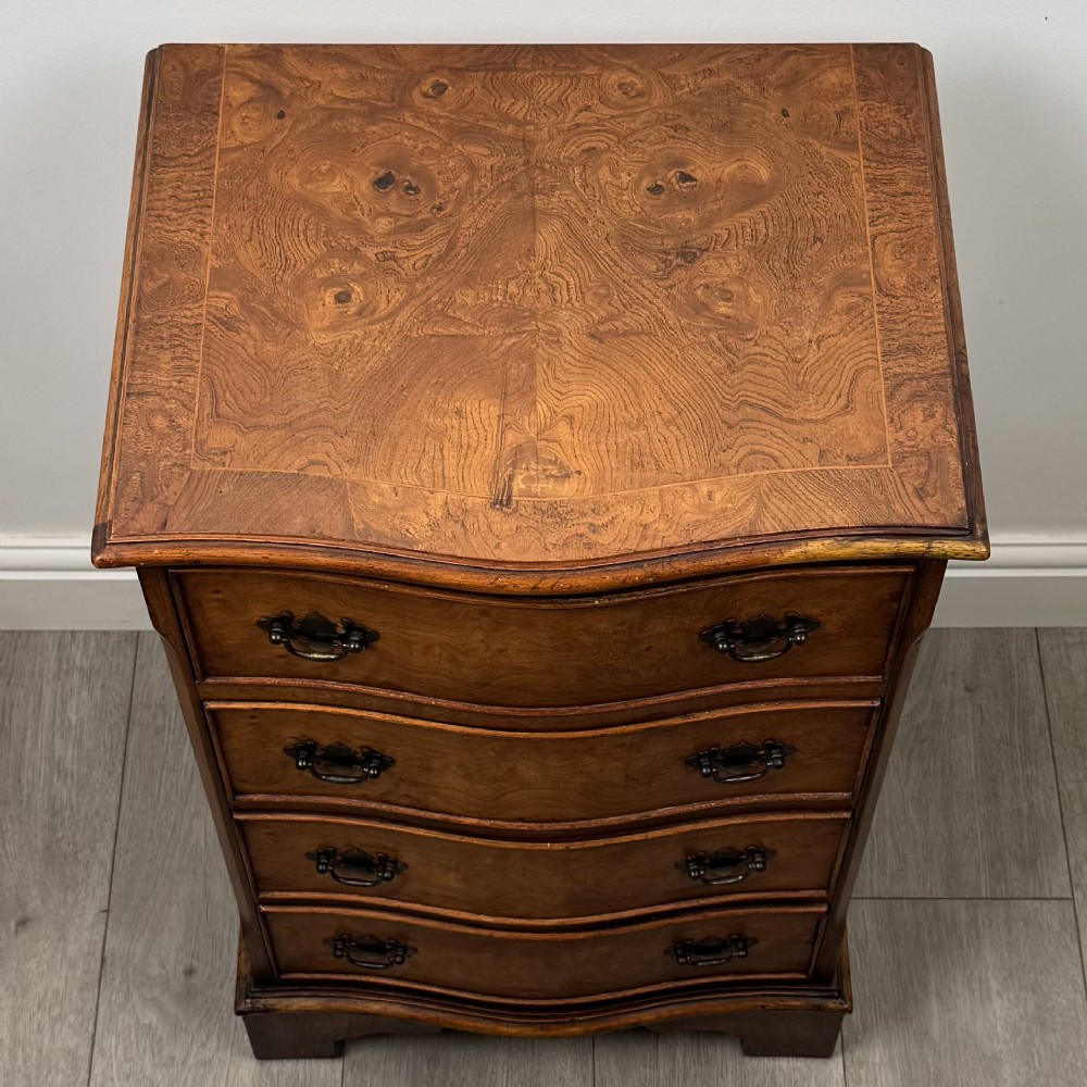 A small 20th-century serpentine chest of drawers with four drawers featuring burr walnut veneer, brass drop handles, and bracket feet. View IX