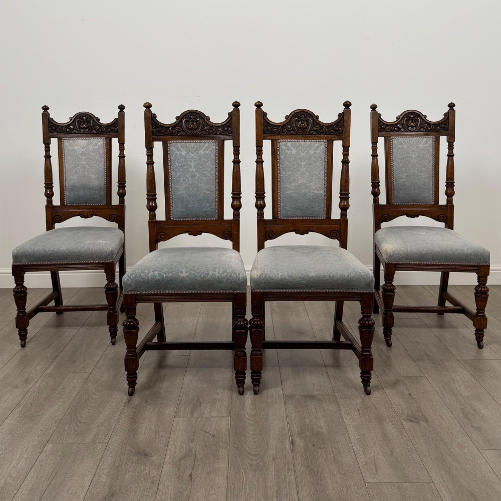 A set of four 19th-century Victorian dark oak dining chairs featuring carved crest rails, turned legs, and light blue patterned fabric upholstery on the seats and backrests.