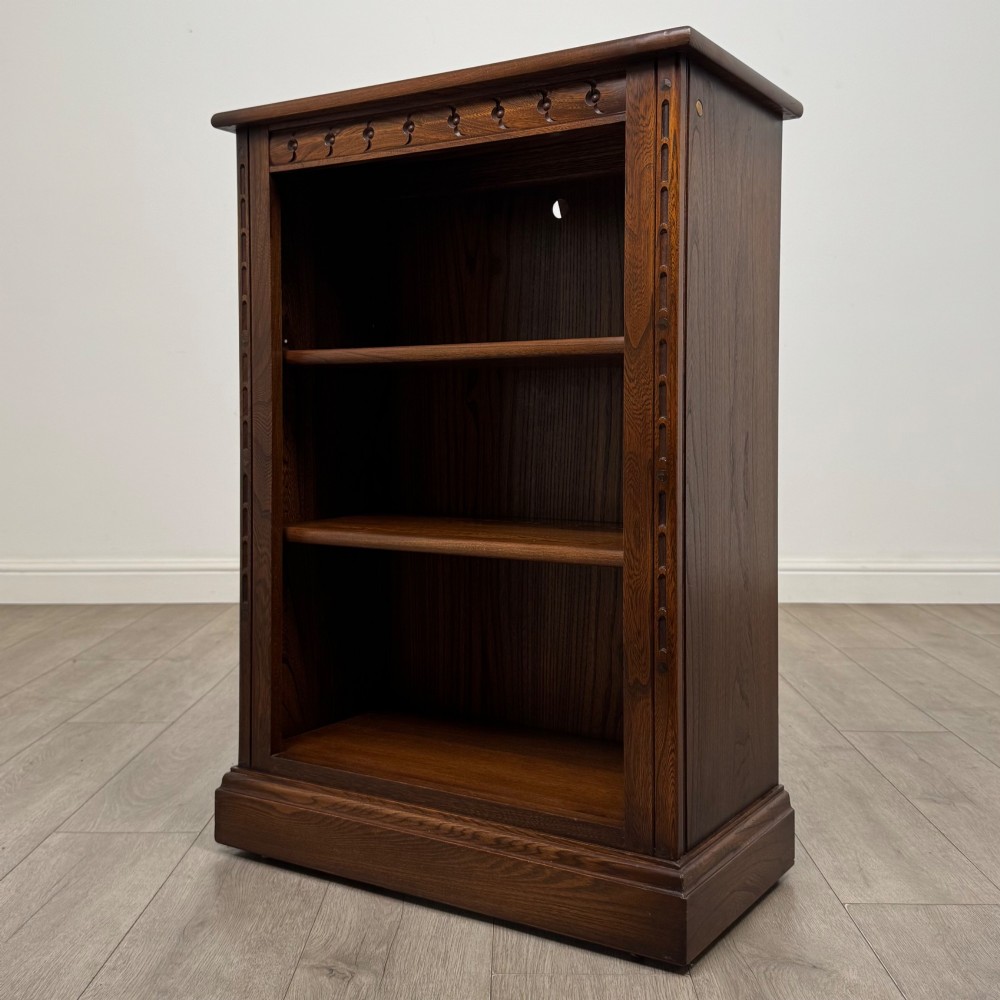 A small vintage dark oak bookcase featuring three open shelves, decorative carved detailing along the frieze and side uprights, and a solid plinth base. View III