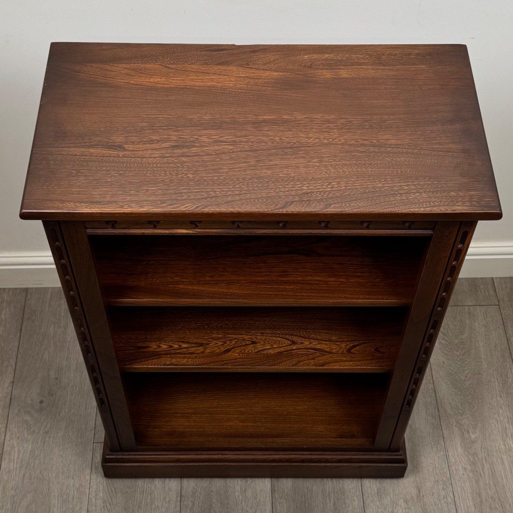 A small vintage dark oak bookcase featuring three open shelves, decorative carved detailing along the frieze and side uprights, and a solid plinth base. View X