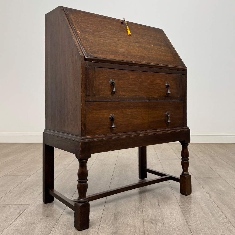 An Edwardian dark oak writing bureau featuring a sloped drop-down lid, two lower drawers with teardrop handles, and turned legs connected by stretchers. View IX