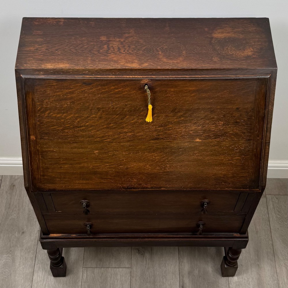 An Edwardian dark oak writing bureau featuring a sloped drop-down lid, two lower drawers with teardrop handles, and turned legs connected by stretchers. View X