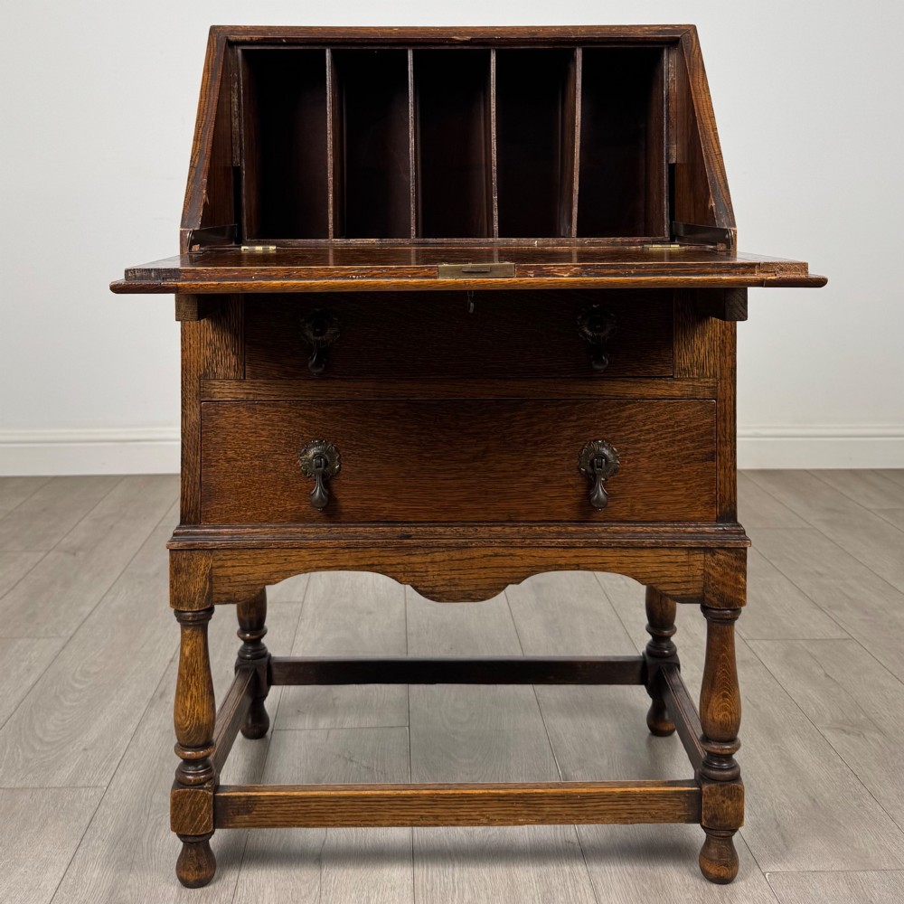 An Edwardian golden oak writing bureau featuring a slant-front writing flap above two drawers with brass teardrop handles, supported on turned legs with a perimeter stretcher. View II
