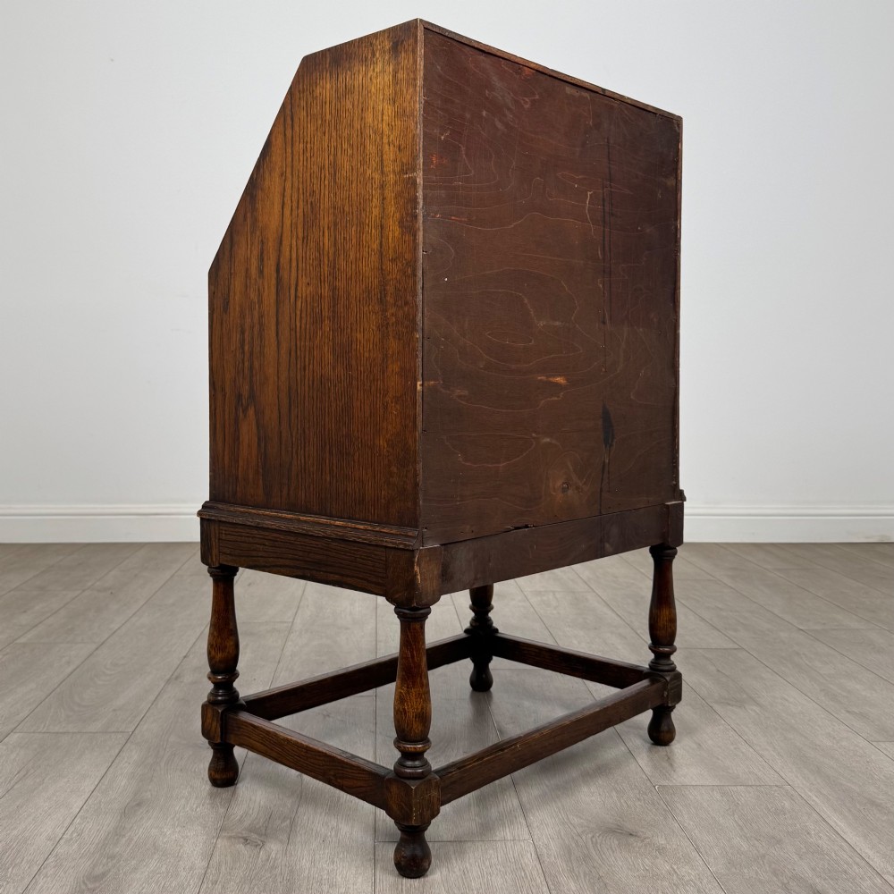 An Edwardian golden oak writing bureau featuring a slant-front writing flap above two drawers with brass teardrop handles, supported on turned legs with a perimeter stretcher. View V
