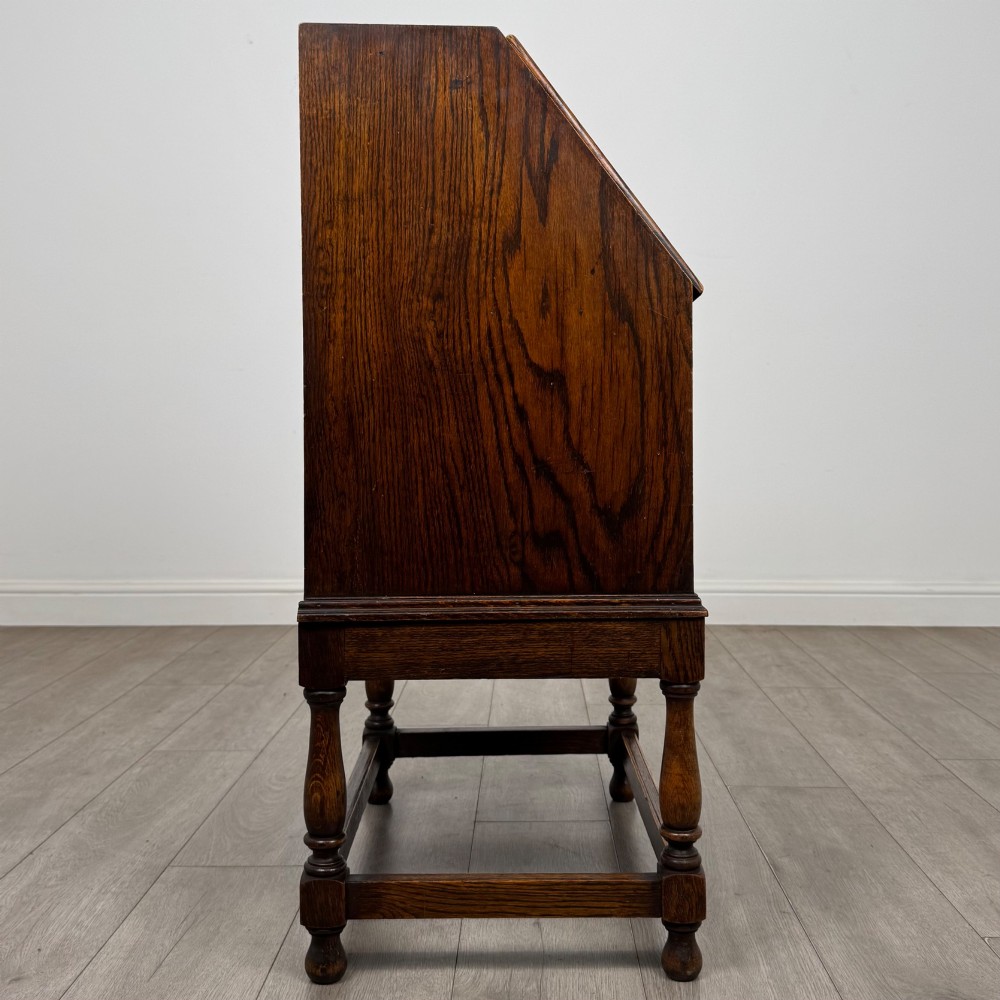 An Edwardian golden oak writing bureau featuring a slant-front writing flap above two drawers with brass teardrop handles, supported on turned legs with a perimeter stretcher. View VIII