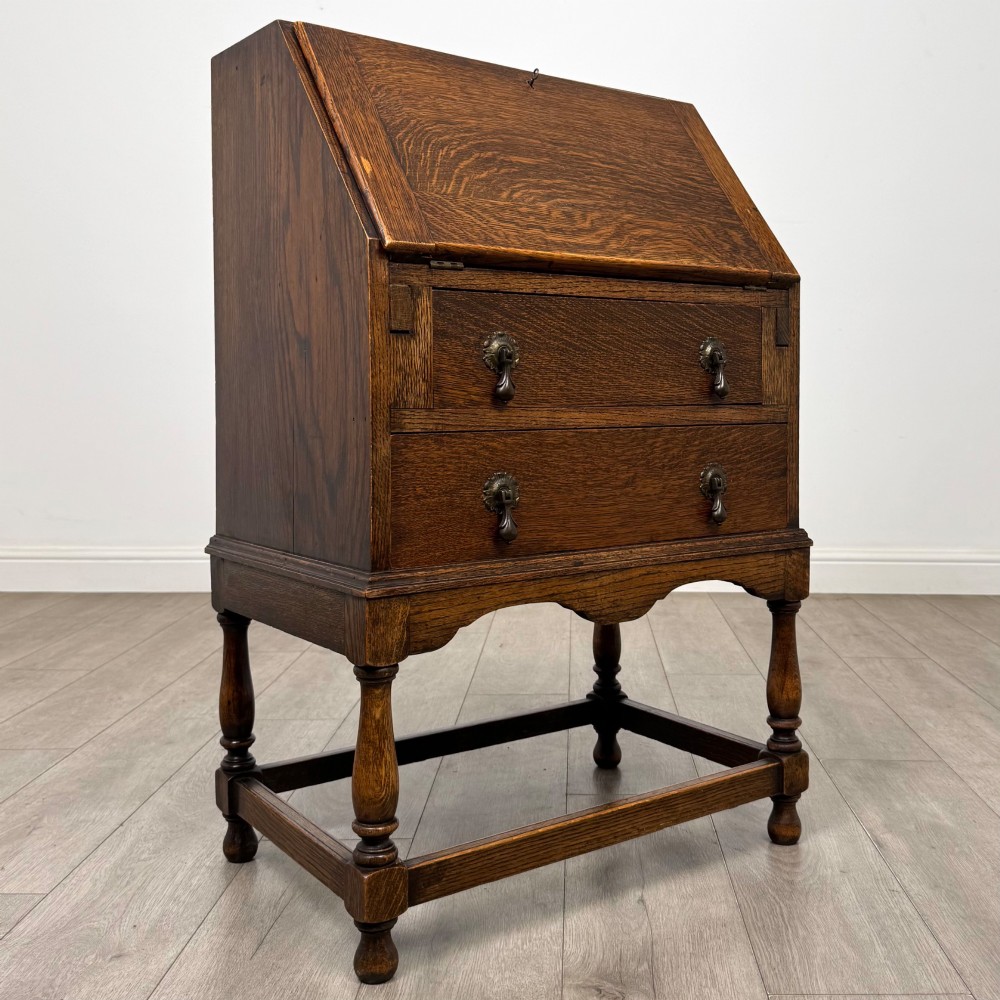 An Edwardian golden oak writing bureau featuring a slant-front writing flap above two drawers with brass teardrop handles, supported on turned legs with a perimeter stretcher. View IX