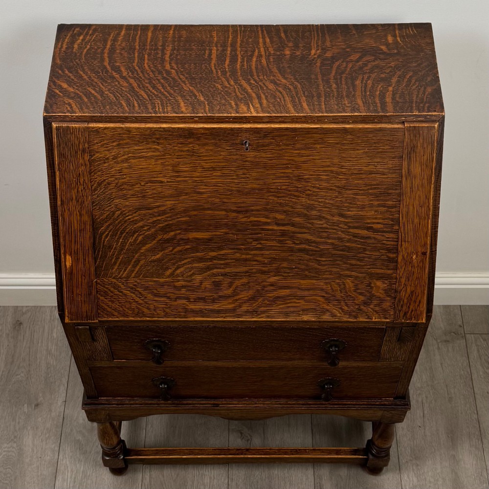 An Edwardian golden oak writing bureau featuring a slant-front writing flap above two drawers with brass teardrop handles, supported on turned legs with a perimeter stretcher. View X