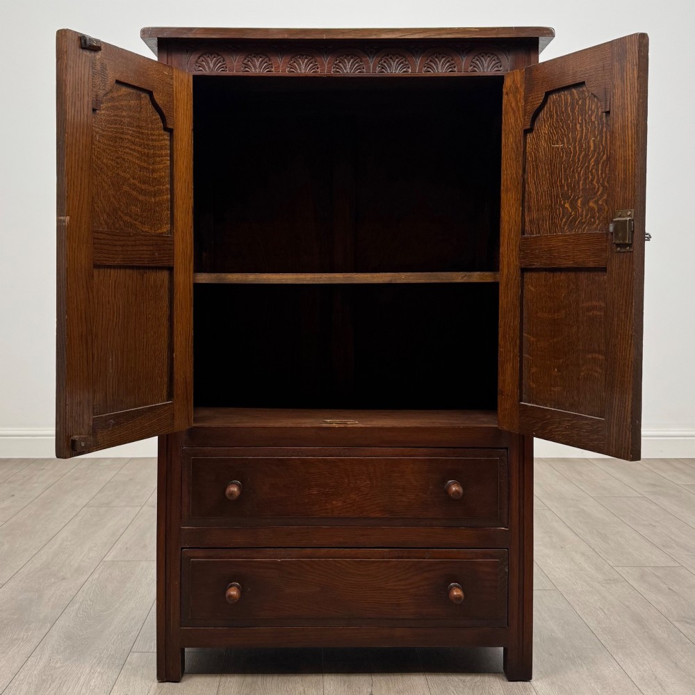 A dark oak cabinet featuring carved top frieze detailing, two arched fielded panel doors with exposed iron hinges, and two lower drawers with turned wooden knobs. View II