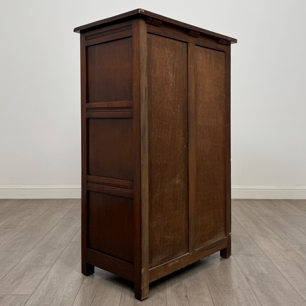 A dark oak cabinet featuring carved top frieze detailing, two arched fielded panel doors with exposed iron hinges, and two lower drawers with turned wooden knobs. View V