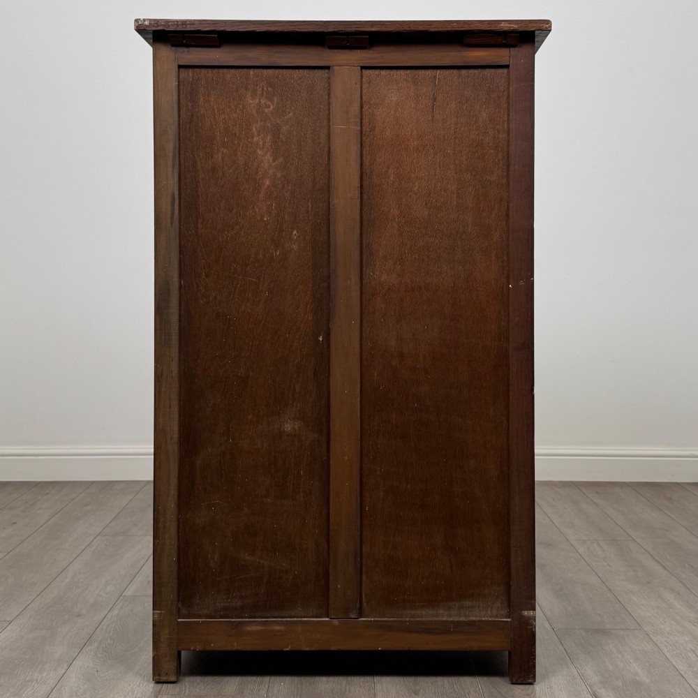 A dark oak cabinet featuring carved top frieze detailing, two arched fielded panel doors with exposed iron hinges, and two lower drawers with turned wooden knobs. View VI