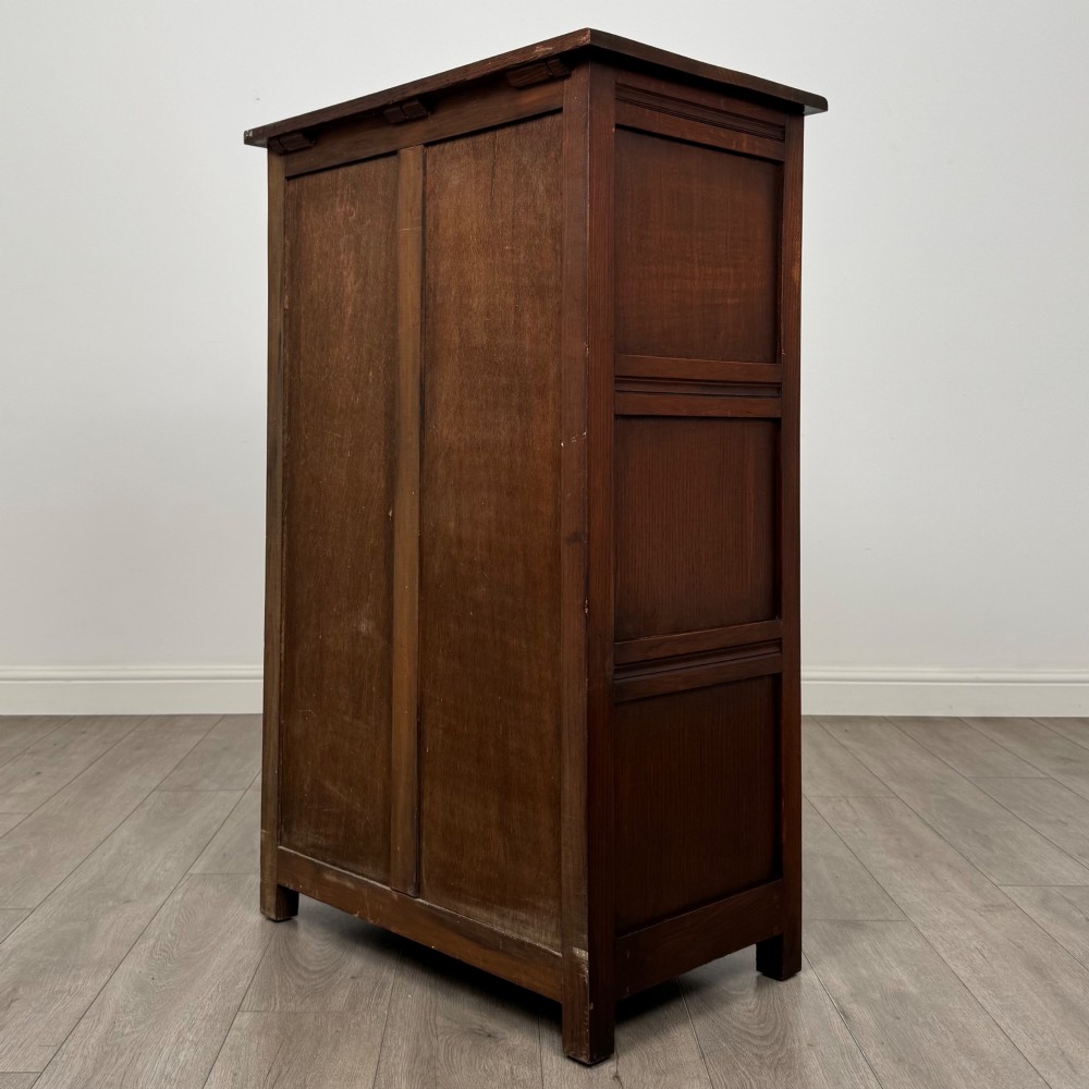 A dark oak cabinet featuring carved top frieze detailing, two arched fielded panel doors with exposed iron hinges, and two lower drawers with turned wooden knobs. View VII