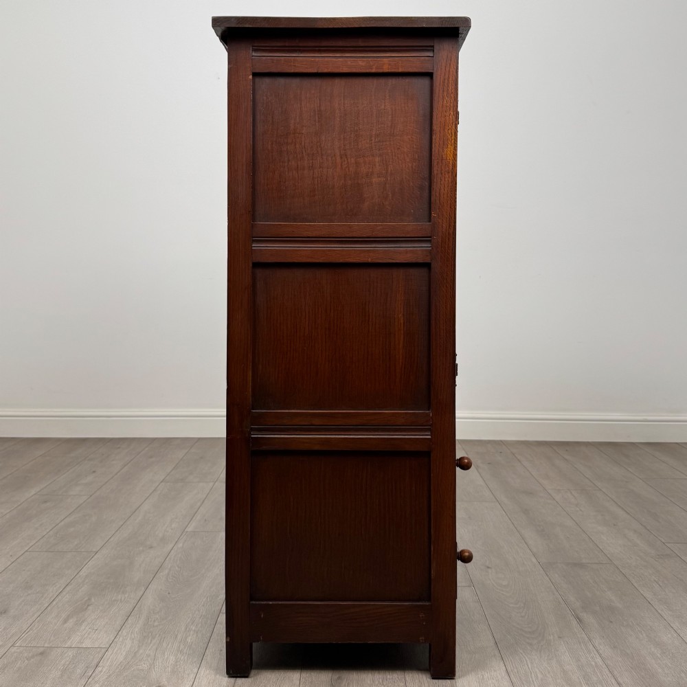 A dark oak cabinet featuring carved top frieze detailing, two arched fielded panel doors with exposed iron hinges, and two lower drawers with turned wooden knobs. View VIII
