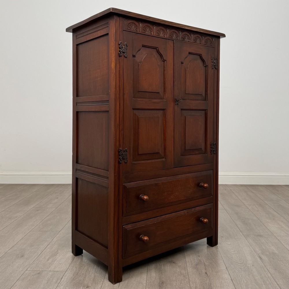 A dark oak cabinet featuring carved top frieze detailing, two arched fielded panel doors with exposed iron hinges, and two lower drawers with turned wooden knobs. View IX