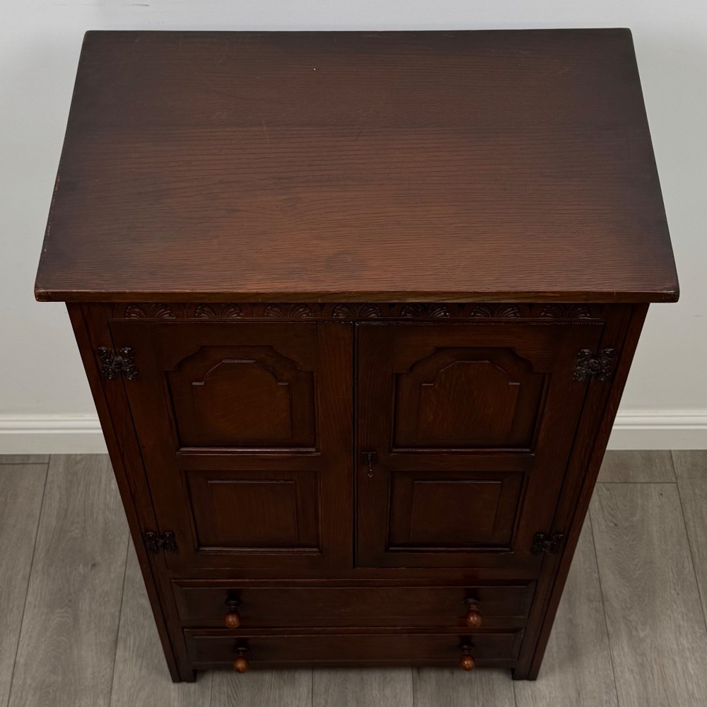 A dark oak cabinet featuring carved top frieze detailing, two arched fielded panel doors with exposed iron hinges, and two lower drawers with turned wooden knobs. View X