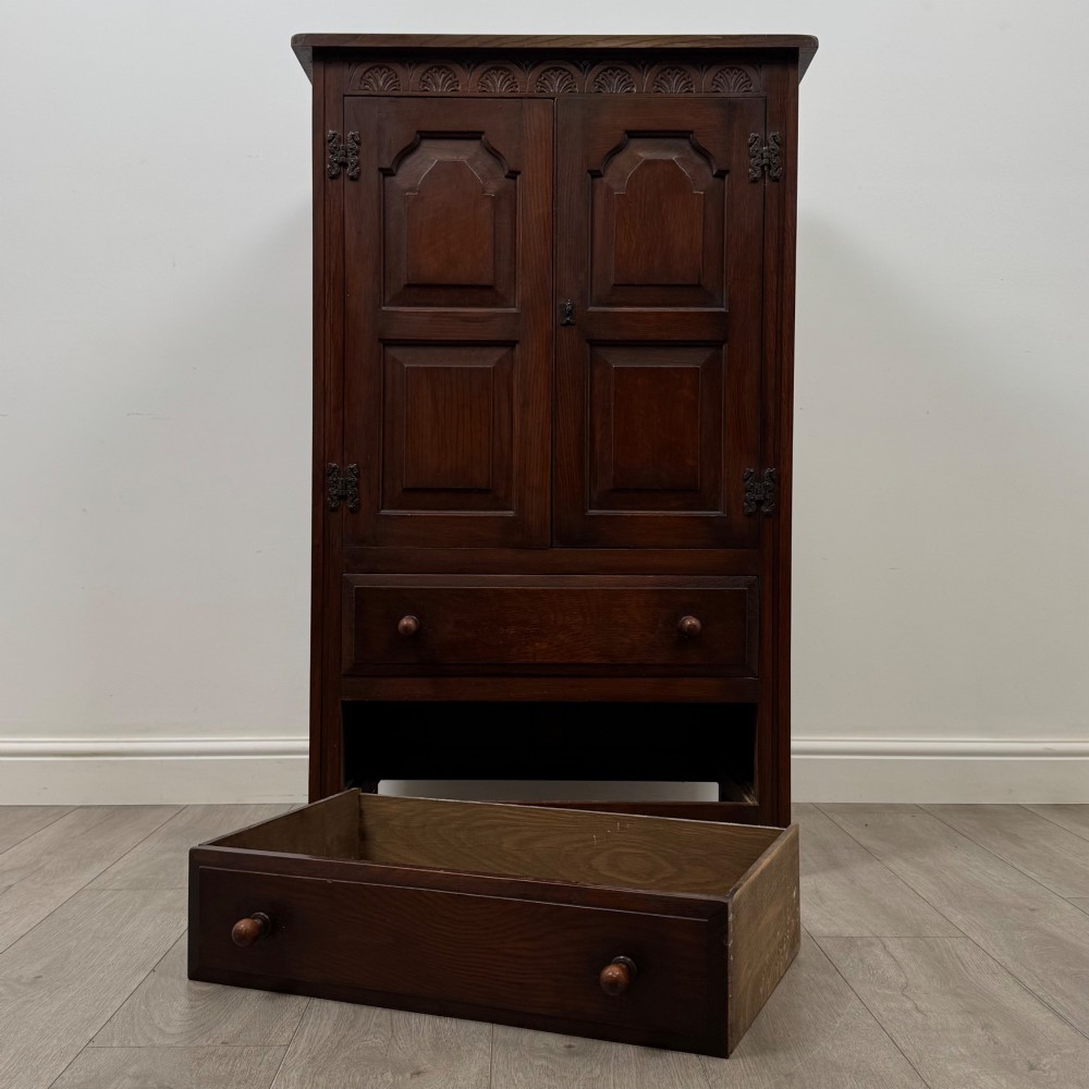 A dark oak cabinet featuring carved top frieze detailing, two arched fielded panel doors with exposed iron hinges, and two lower drawers with turned wooden knobs. View XI