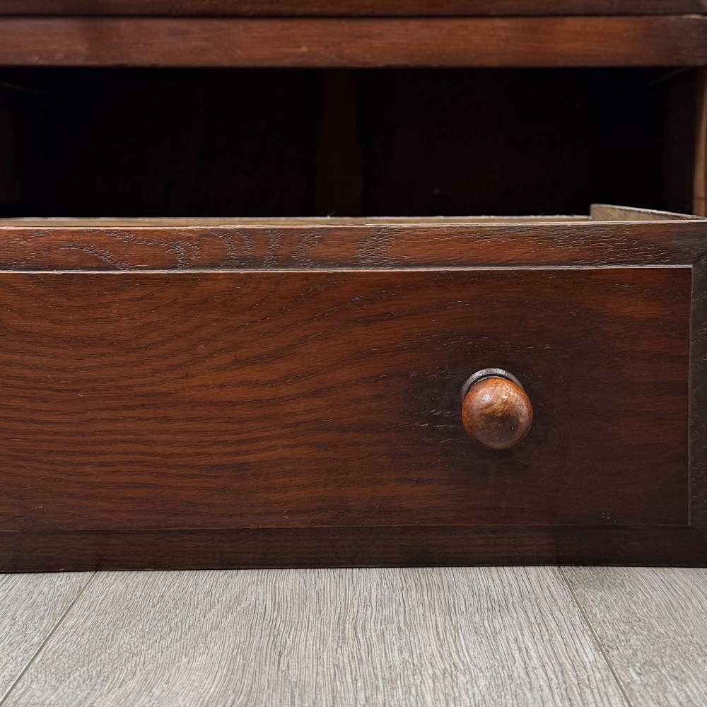 A dark oak cabinet featuring carved top frieze detailing, two arched fielded panel doors with exposed iron hinges, and two lower drawers with turned wooden knobs. View XII