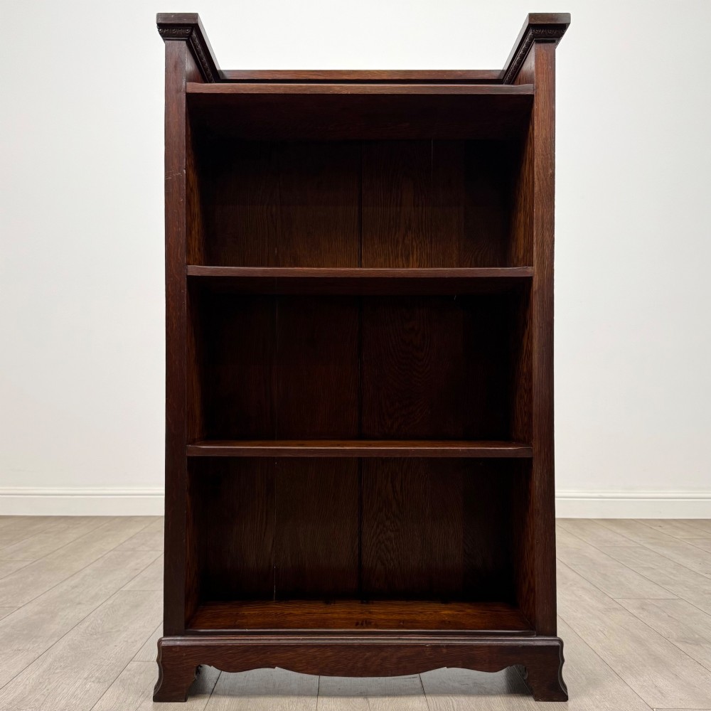 An Edwardian open oak bookcase featuring three interior fixed shelves, dark timber finish, carved moulding on top uprights, and a shaped plinth base. View II