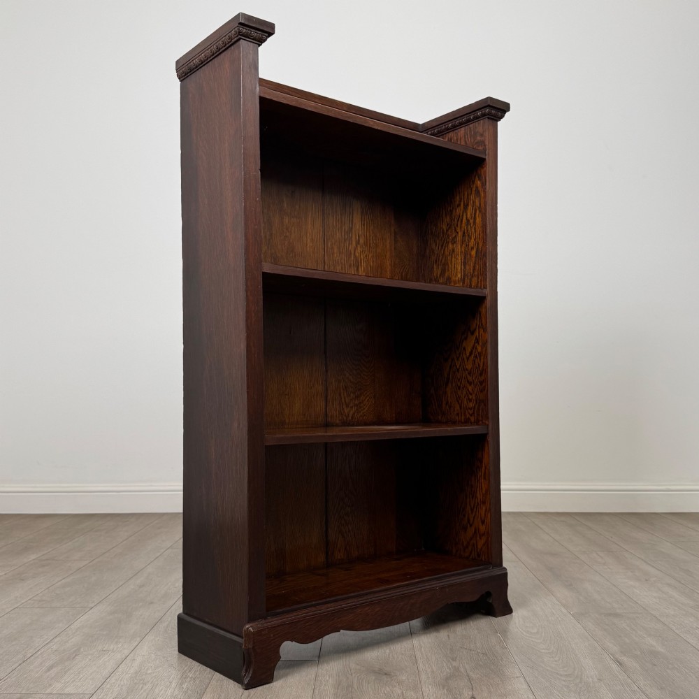 An Edwardian open oak bookcase featuring three interior fixed shelves, dark timber finish, carved moulding on top uprights, and a shaped plinth base. View IX
