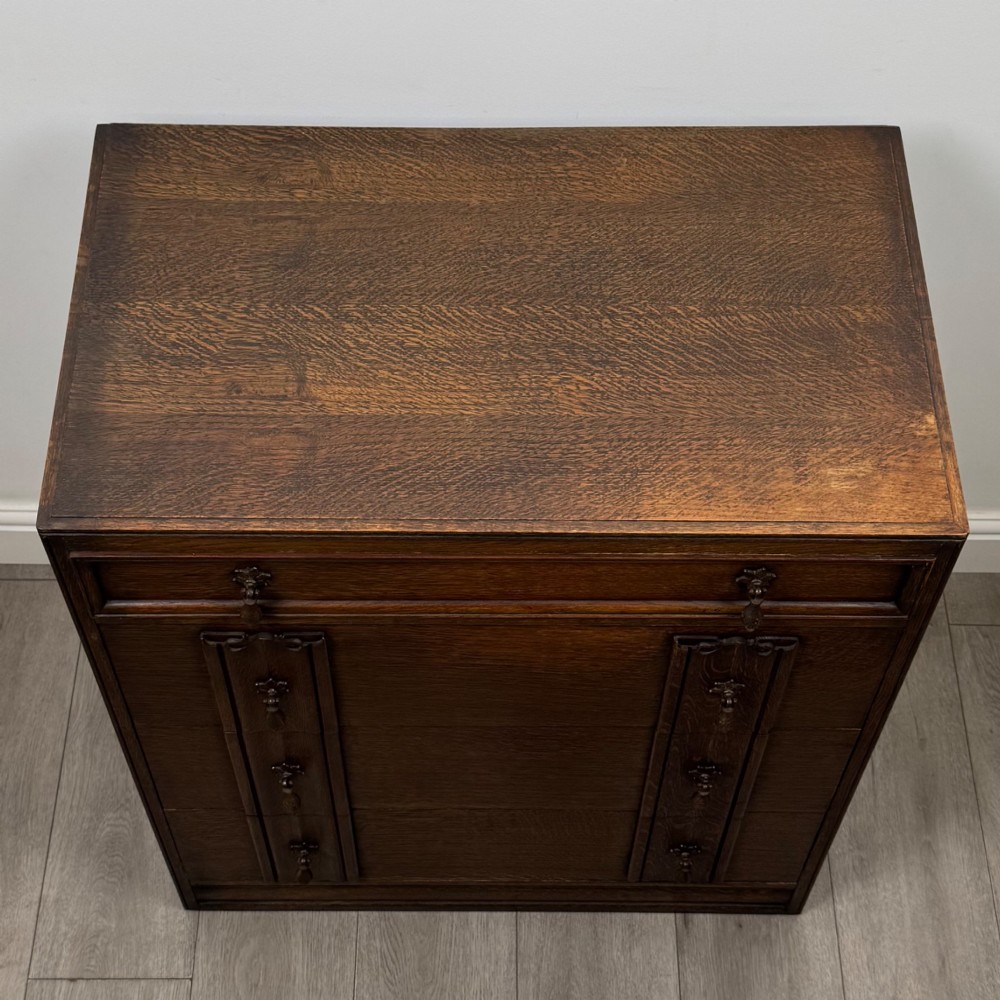 A dark oak chest of drawers featuring carved vertical linenfold panels and metallic drop handles. View IX