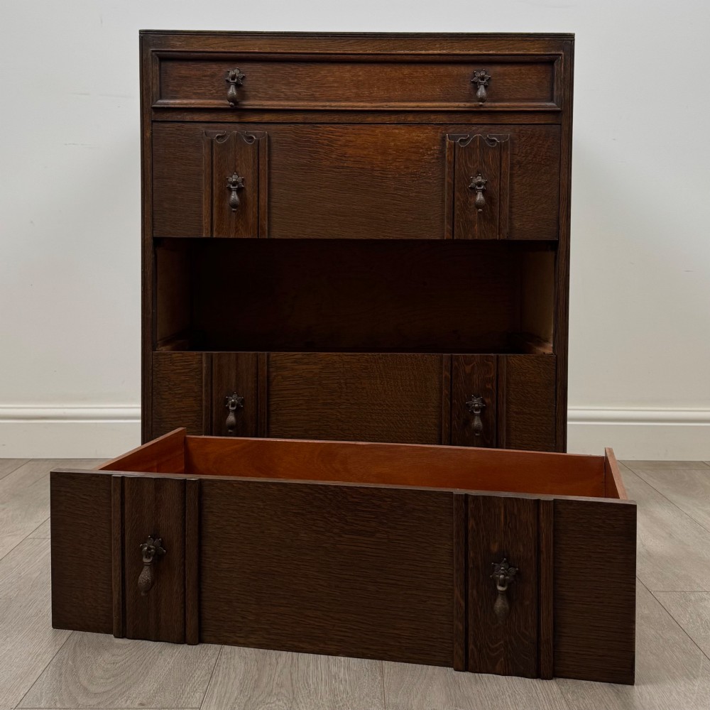 A dark oak chest of drawers featuring carved vertical linenfold panels and metallic drop handles. View X