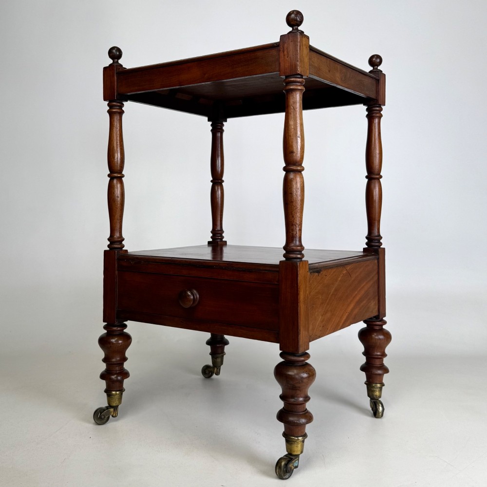 A Victorian mahogany two-tiered stand featuring turned corner supports, top finials, a lower single drawer with a round wooden knob, and brass castors. View II