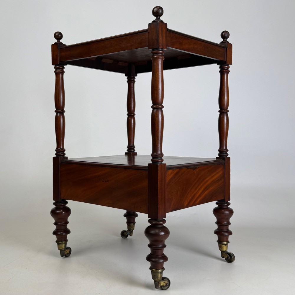 A Victorian mahogany two-tiered stand featuring turned corner supports, top finials, a lower single drawer with a round wooden knob, and brass castors. View VI