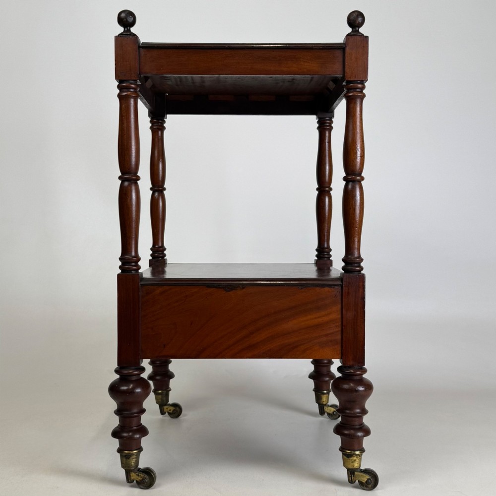 A Victorian mahogany two-tiered stand featuring turned corner supports, top finials, a lower single drawer with a round wooden knob, and brass castors. View VII