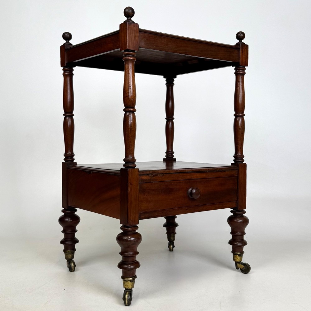 A Victorian mahogany two-tiered stand featuring turned corner supports, top finials, a lower single drawer with a round wooden knob, and brass castors. View VIII