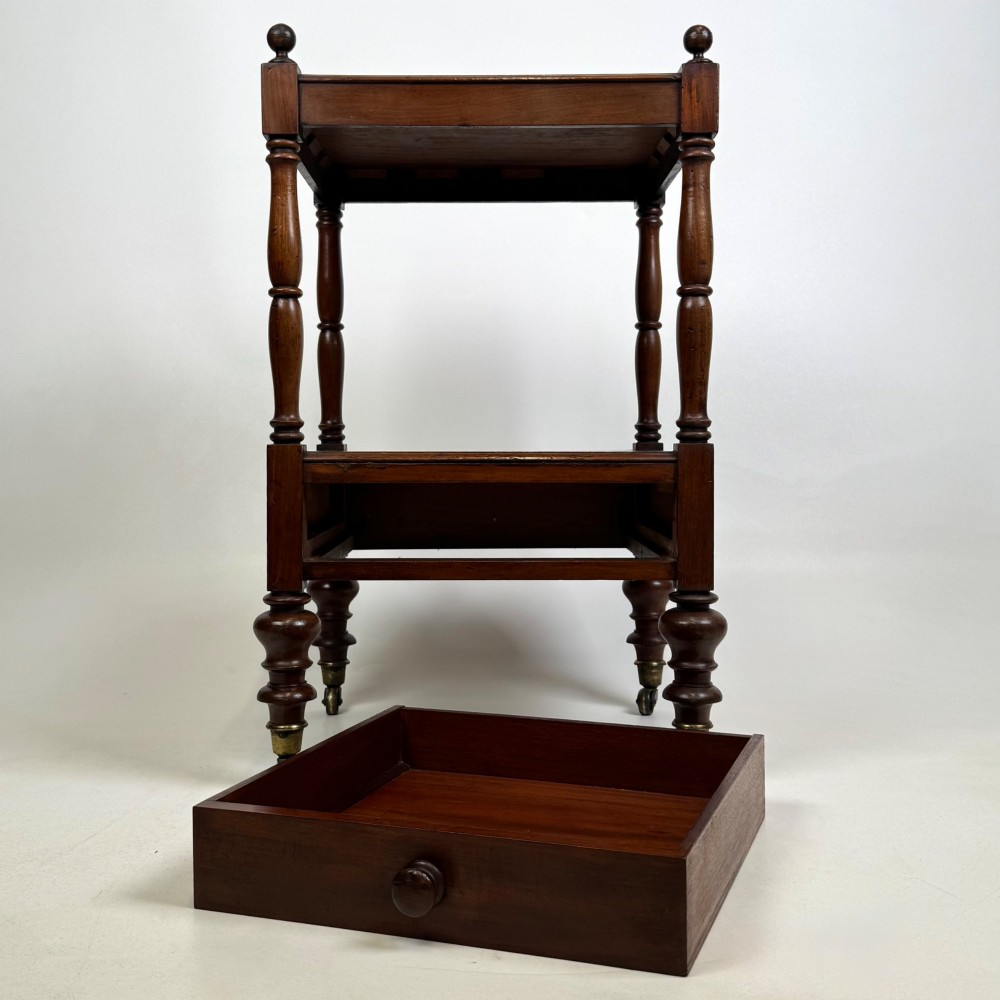 A Victorian mahogany two-tiered stand featuring turned corner supports, top finials, a lower single drawer with a round wooden knob, and brass castors. View IX