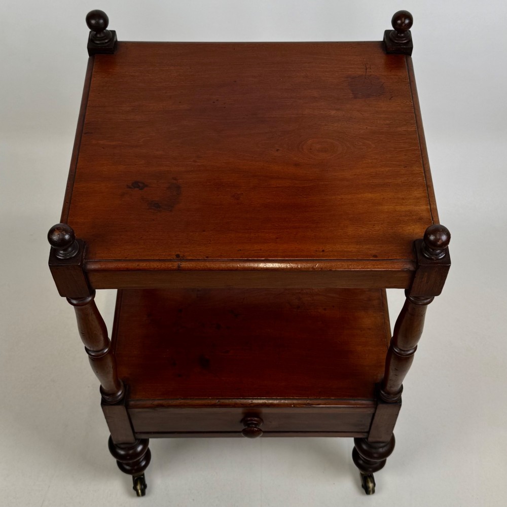 A Victorian mahogany two-tiered stand featuring turned corner supports, top finials, a lower single drawer with a round wooden knob, and brass castors. View X