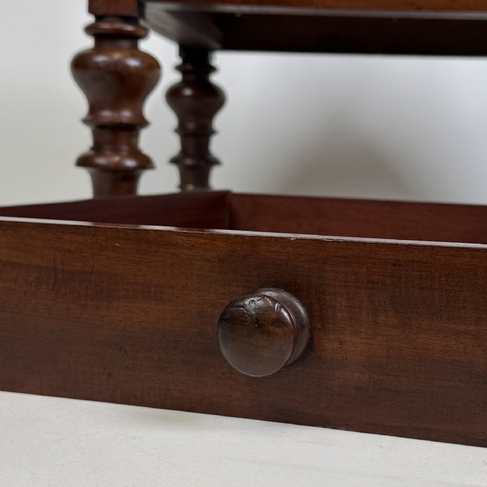 A Victorian mahogany two-tiered stand featuring turned corner supports, top finials, a lower single drawer with a round wooden knob, and brass castors. View XI