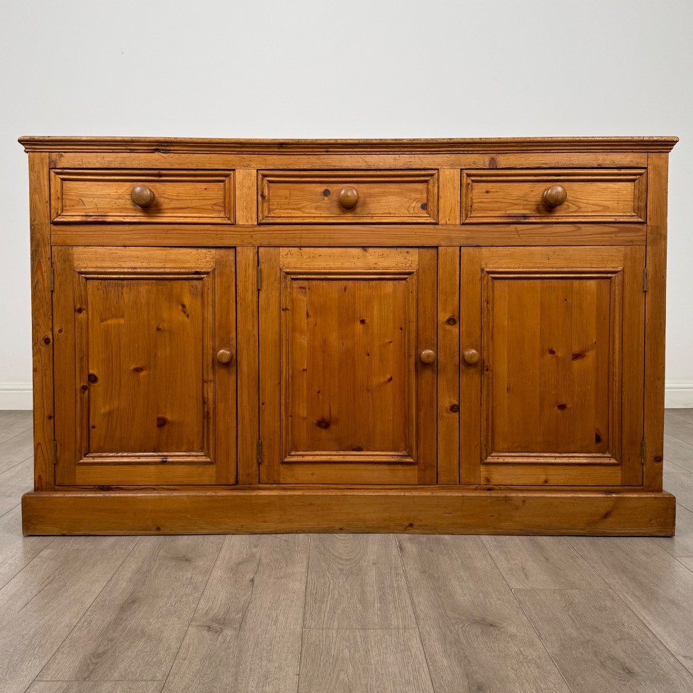 A vintage pine farmhouse sideboard featuring three frieze drawers above three panelled cupboard doors with turned wooden knobs on a plinth base.
