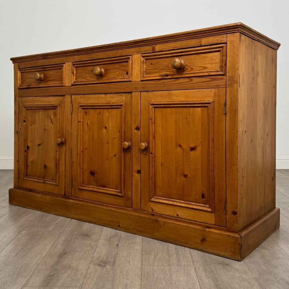 A vintage pine farmhouse sideboard featuring three frieze drawers above three panelled cupboard doors with turned wooden knobs on a plinth base. View III