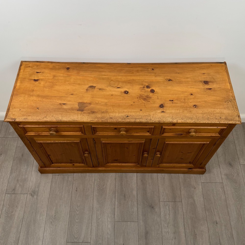 A vintage pine farmhouse sideboard featuring three frieze drawers above three panelled cupboard doors with turned wooden knobs on a plinth base. View X