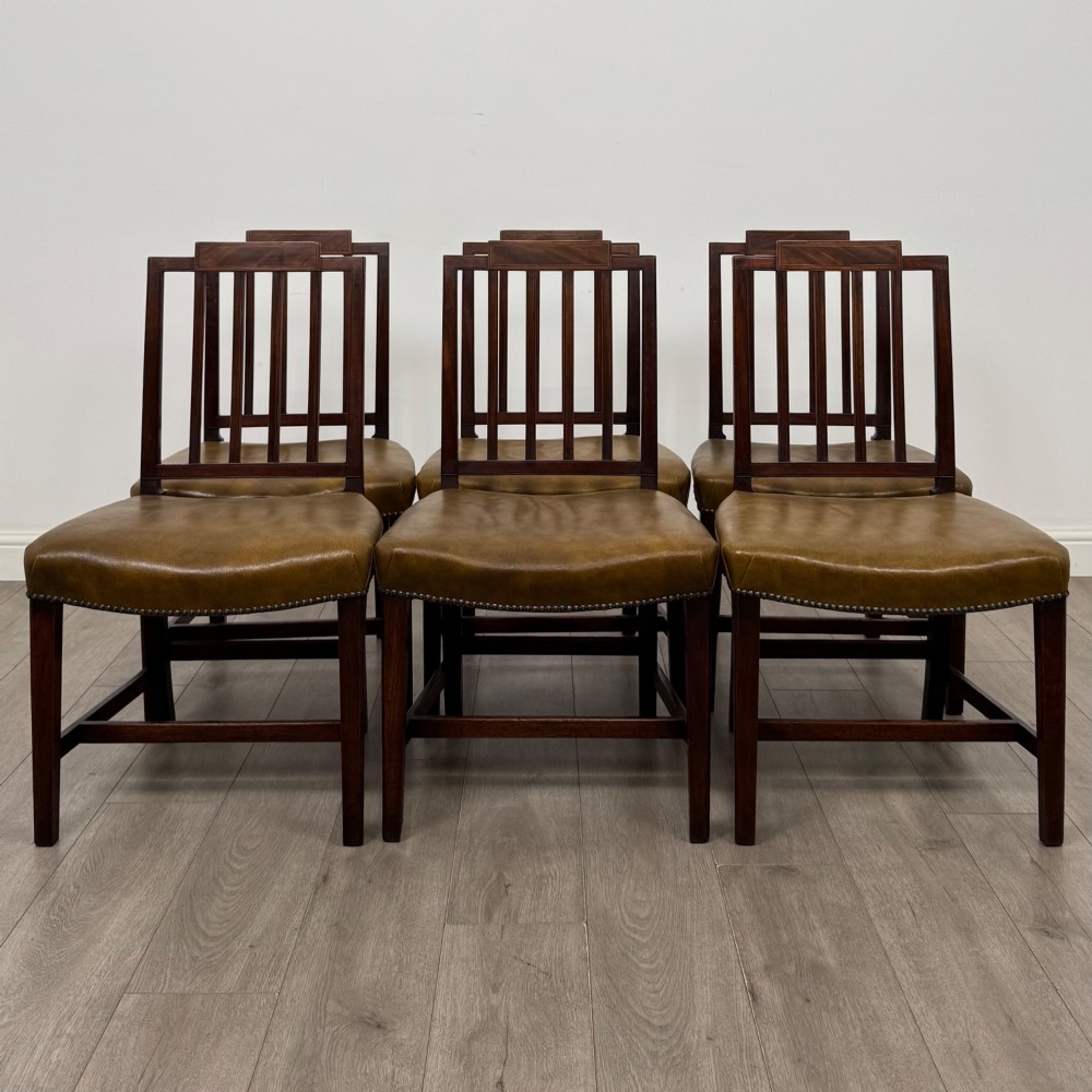 A set of six antique mahogany dining chairs featuring vertical slat backs, square legs, and olive green leather seats finished with brass studs.